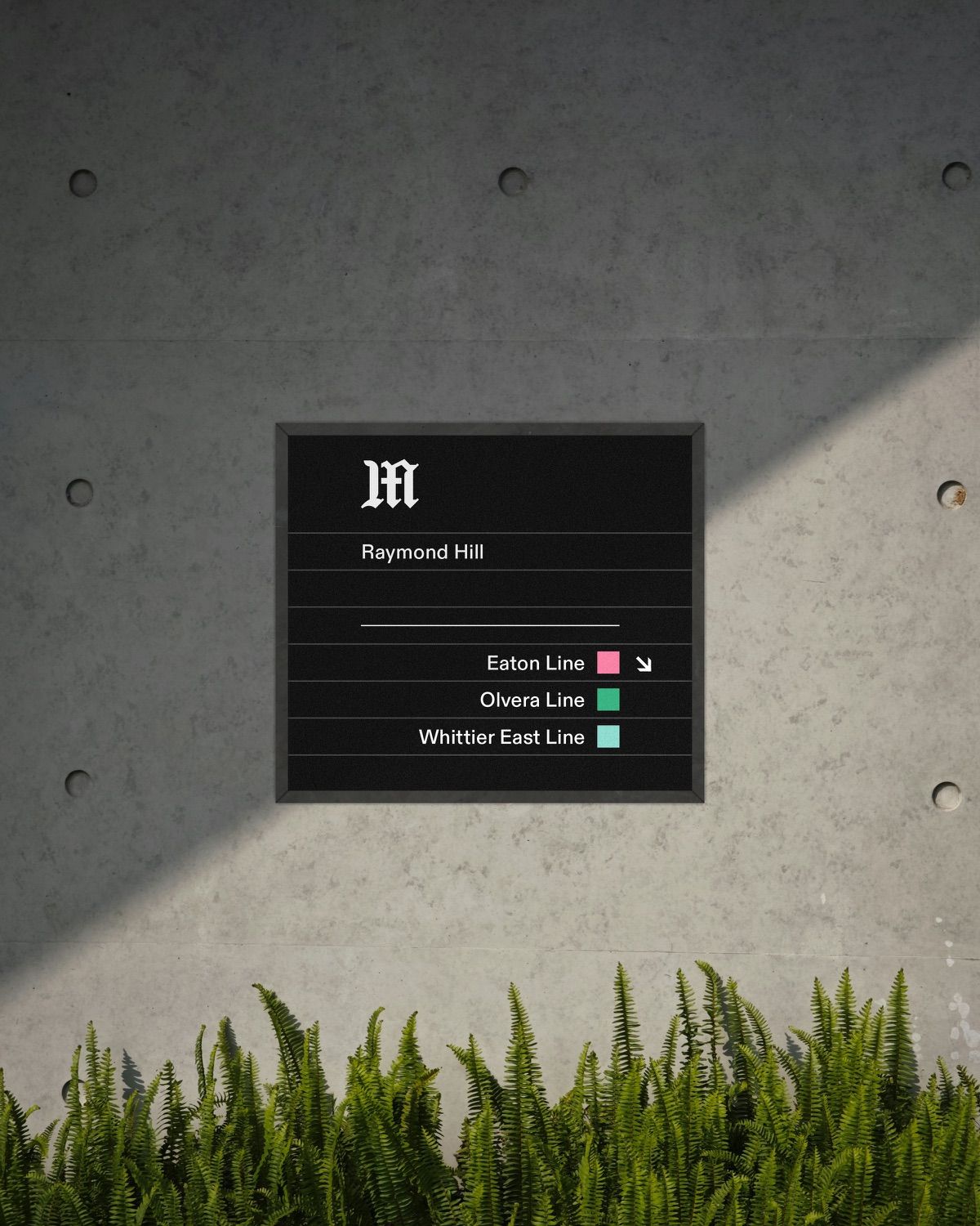 A wall-mounted station sign for Raymond Hill on a concrete wall above grass.