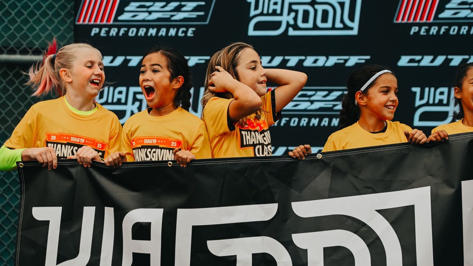 Young players in Via Footy shirts cheering behind a Via Footy banner.