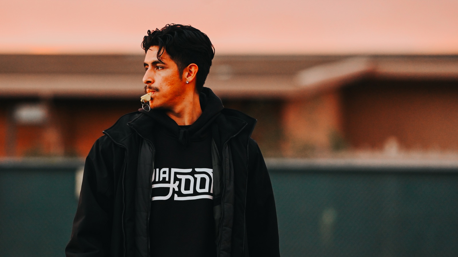 A coach in a Via Footy hoodie with a whistle, at dusk.