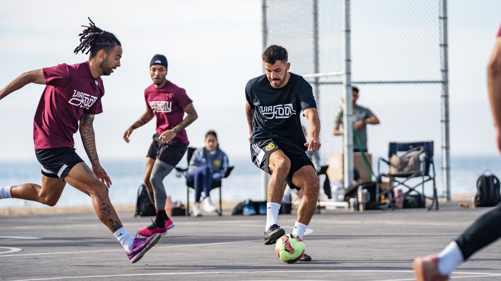 Players in Via Footy kit contesting the ball on an outdoor court by the sea.