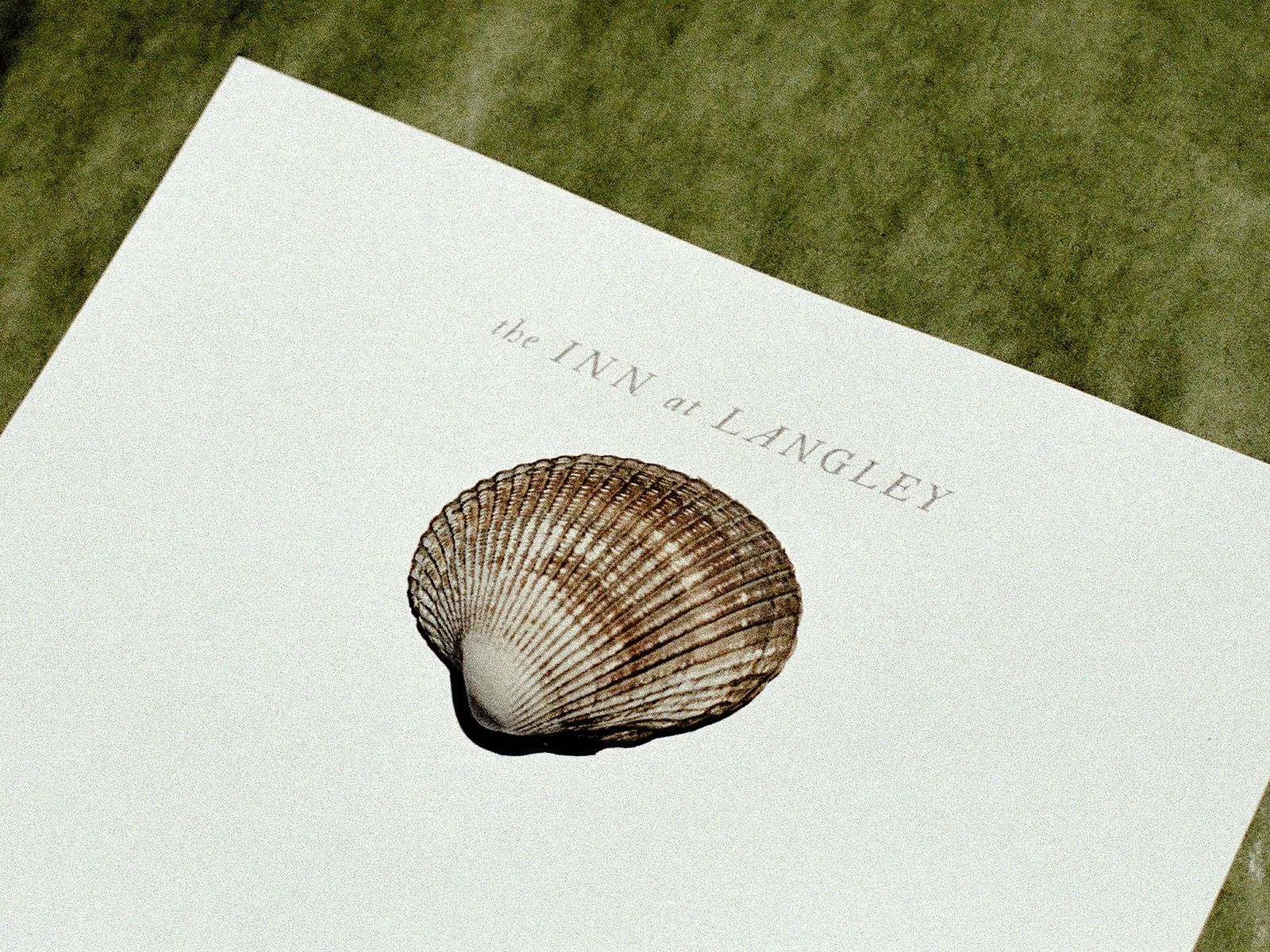 Pale letterhead printed “the Inn at Langley” with a cockle shell resting on it, laid on green fabric.